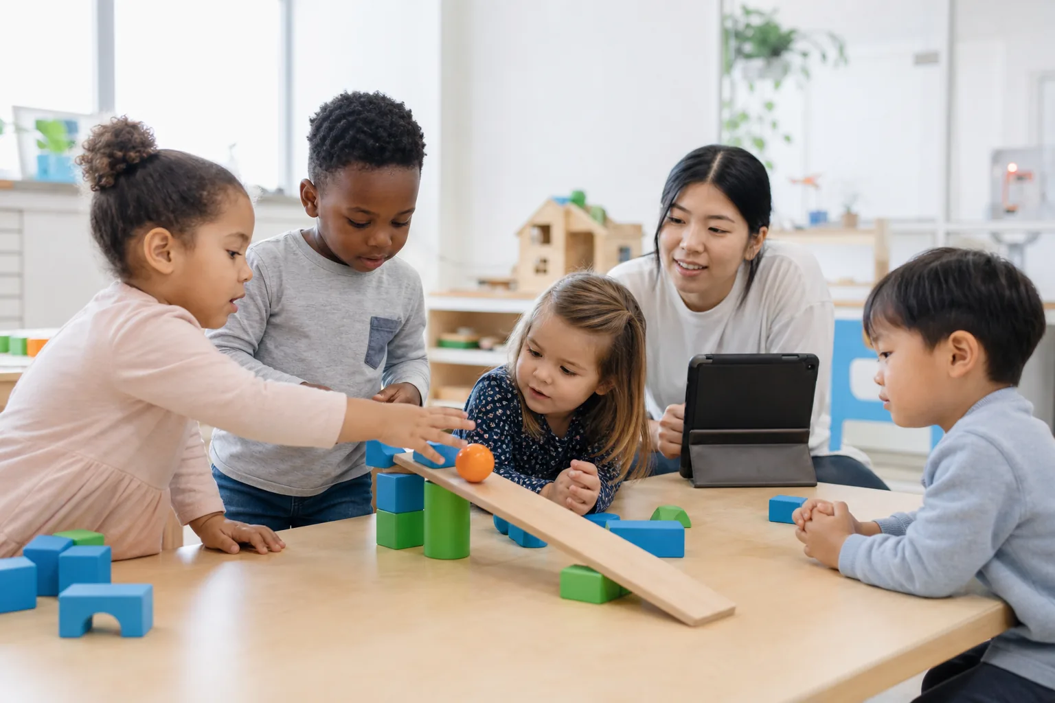 Preschoolers building a ramp with colorful blocks in a STEM activity while a teacher observes nearby