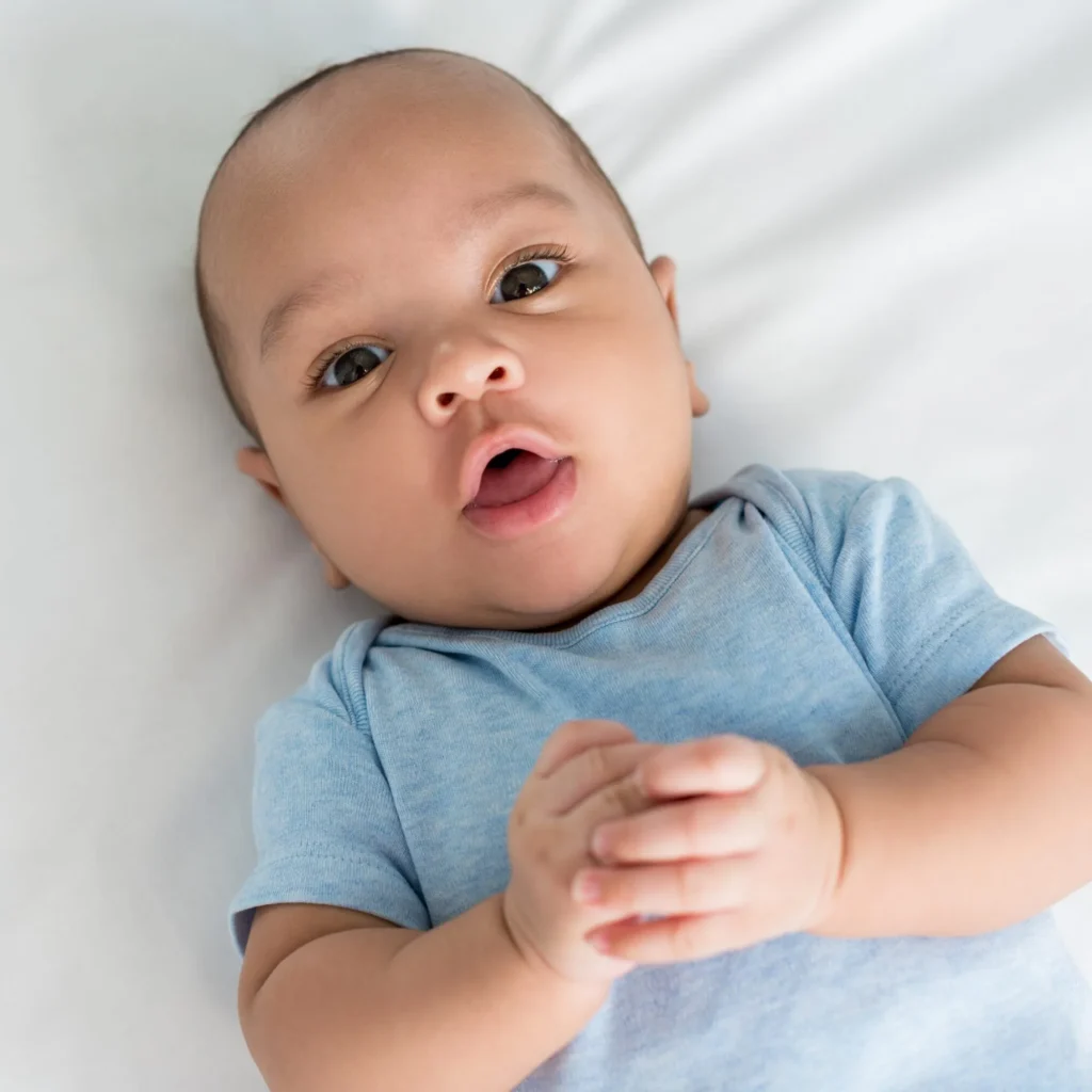 Stock photo of a baby in a light blue bodysuit lying on a white sheet, hands clasped