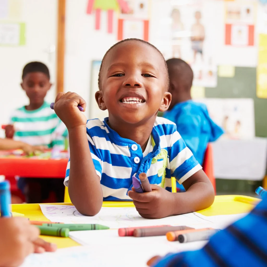Smiling young boy in a striped shirt holding crayons at a classroom table with other children