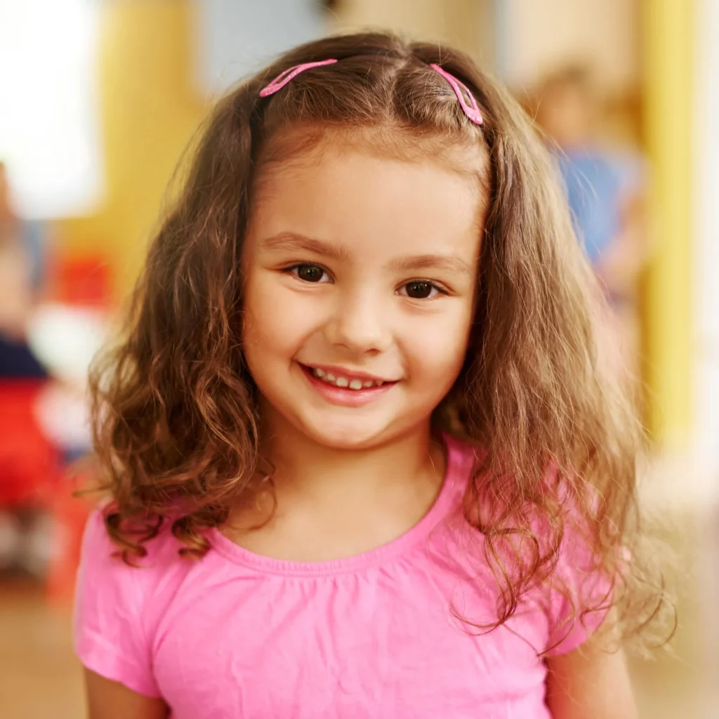 Smiling young girl with curly brown hair, pink hair clips and a pink shirt indoors