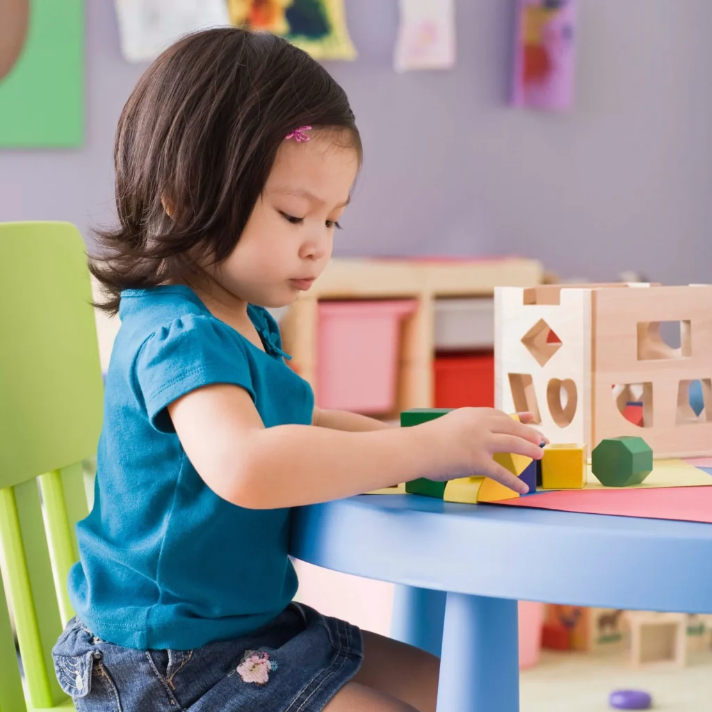 Toddler girl in a teal shirt playing with wooden shape blocks at a blue table